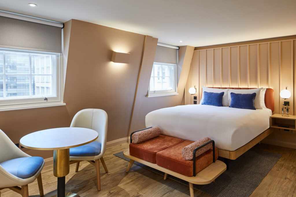 A master Cannon London room with a white bed and navy pillows, a rust-colored bench at its foot, and a small round table with two chairs by a window.