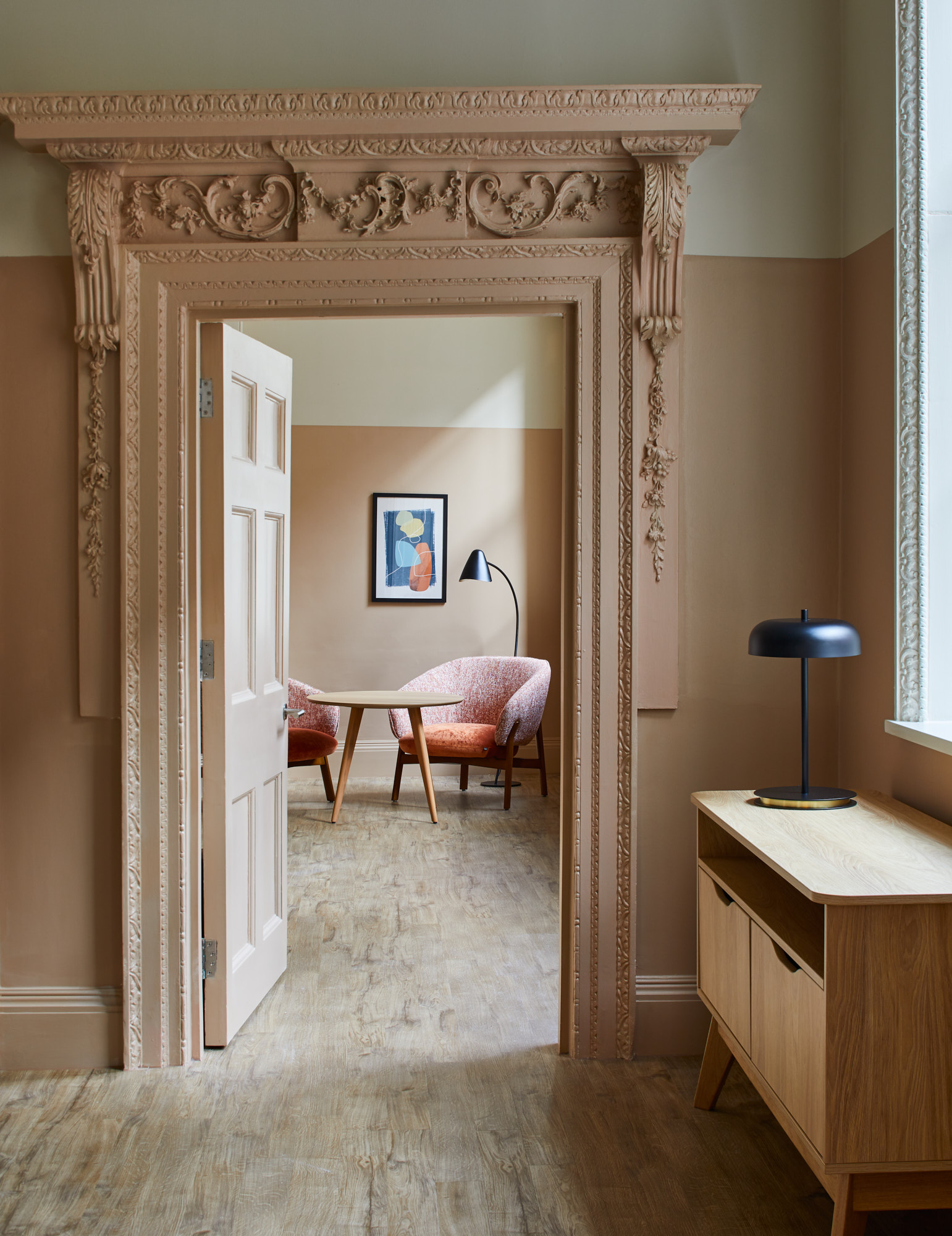 Ornate doorway with intricate mouldings leading to a bright sitting area with pink armchairs and a round table.