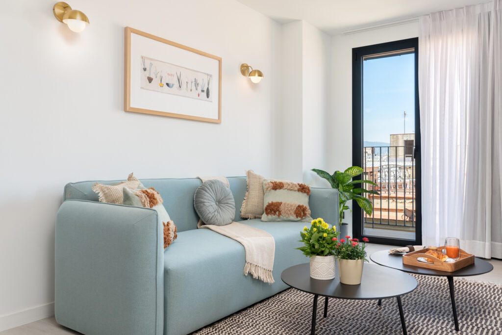 Modern studio at master La Rambla featuring a light blue sofa, indoor plants, and access to a private balcony overlooking the city.