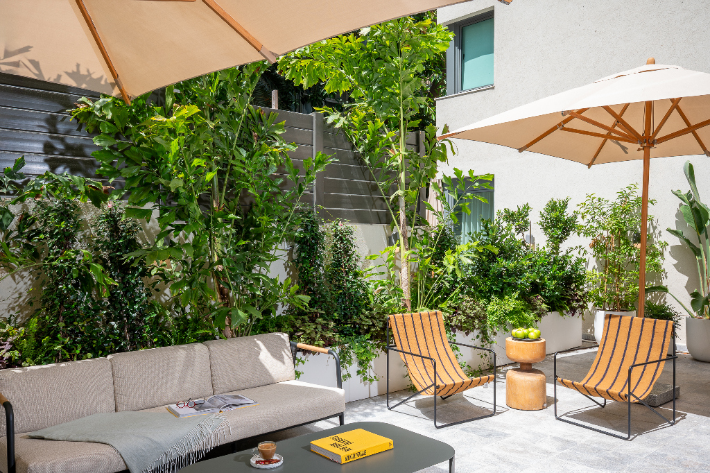 Cozy terrace lounge area at master Mazeh with a cushioned outdoor sofa, striped chairs, greenery-filled planters, and sun umbrellas casting shade across the tranquil space.