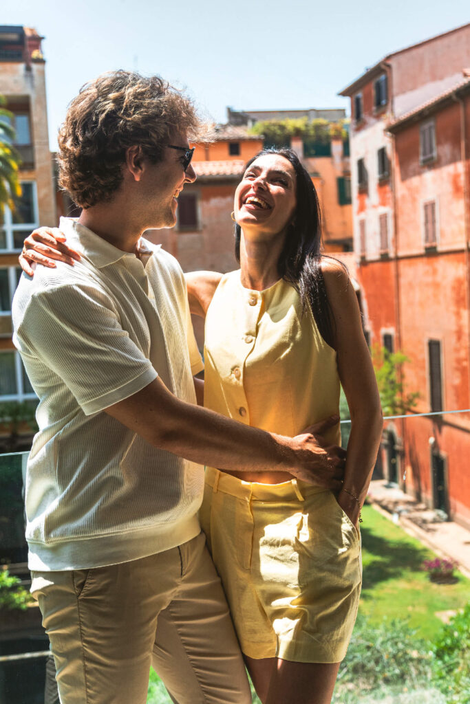 Happy couple smiling and embracing on a sunny balcony with colorful old buildings in the background.