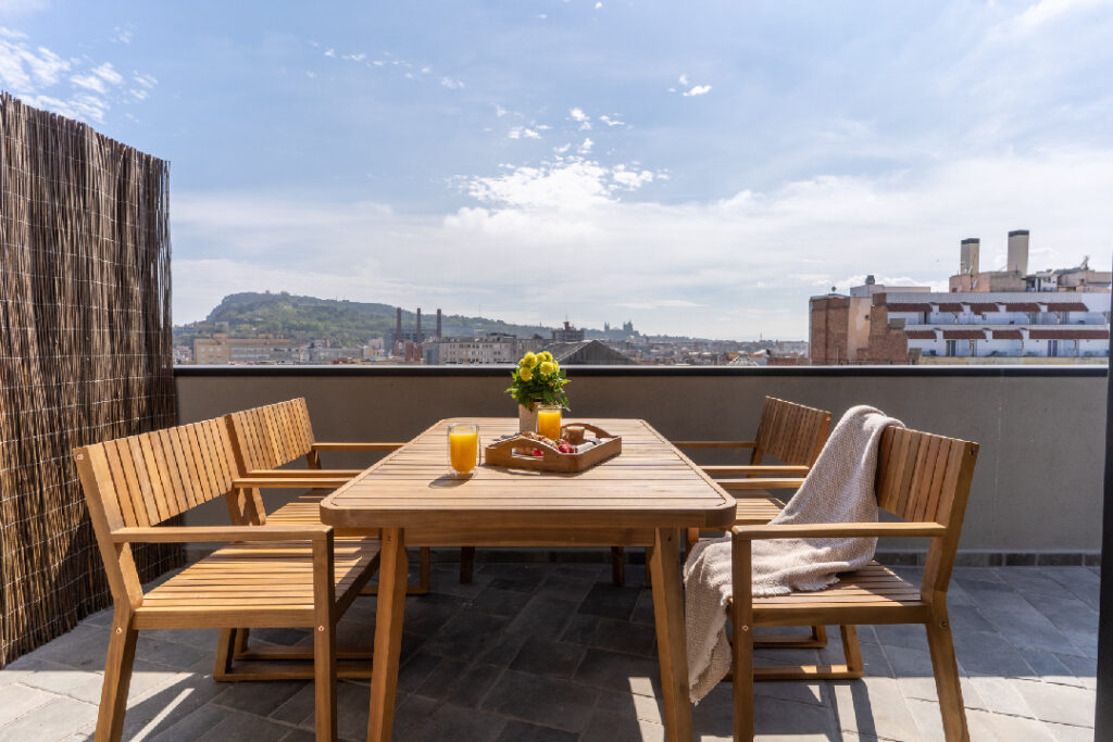 Spacious private penthouse terrace with wooden dining furniture, fresh juice, flowers, and panoramic views over Barcelona’s skyline.