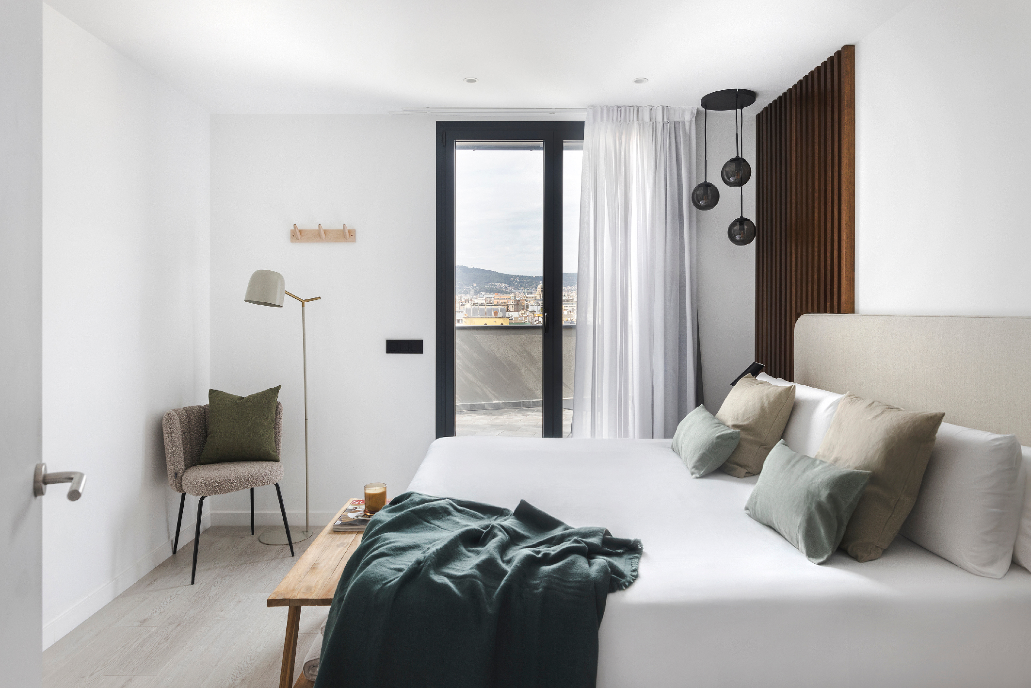 Bright penthouse bedroom with king-size bed, green pillows, cozy seating corner, and floor-to-ceiling doors opening to a terrace overlooking Barcelona.