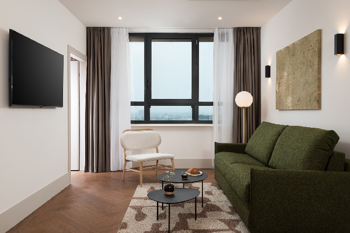 Living room in master Wola two bedroom apartment with green sofa, seating area, wall-mounted TV, and large window.