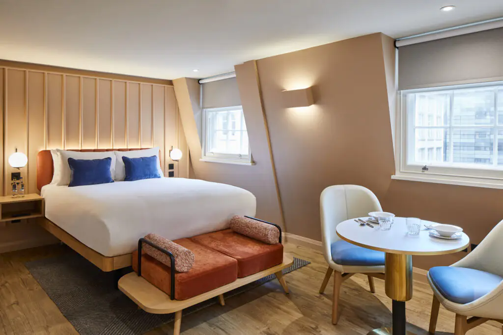 Bright studio at master Cannon London featuring a comfortable bed with blue accent pillows, a compact dining area, and large windows providing natural light.