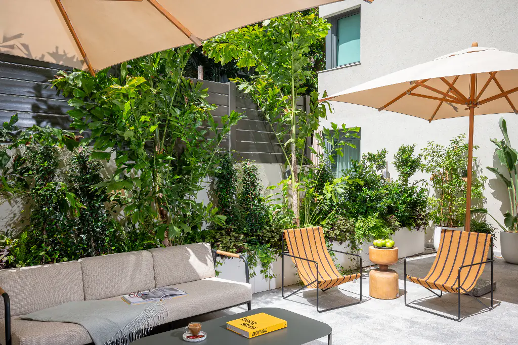 Cozy terrace lounge area at master Mazeh with a cushioned outdoor sofa, striped chairs, greenery-filled planters, and sun umbrellas casting shade across the tranquil space.