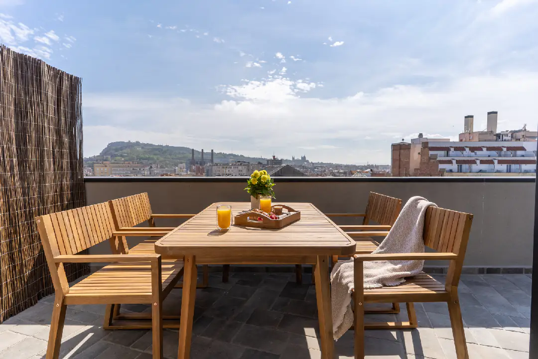 Spacious private penthouse terrace with wooden dining furniture, fresh juice, flowers, and panoramic views over Barcelona’s skyline.
